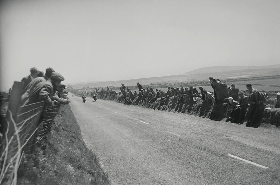 Three riders race towards Creg-ny-Baa seen from behind looking down the road