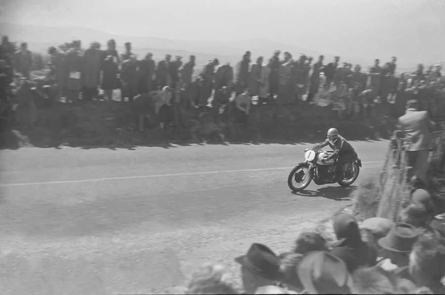 Geoff Duke (#1) from the side as he passes through the crowds on his motorcyle at speed